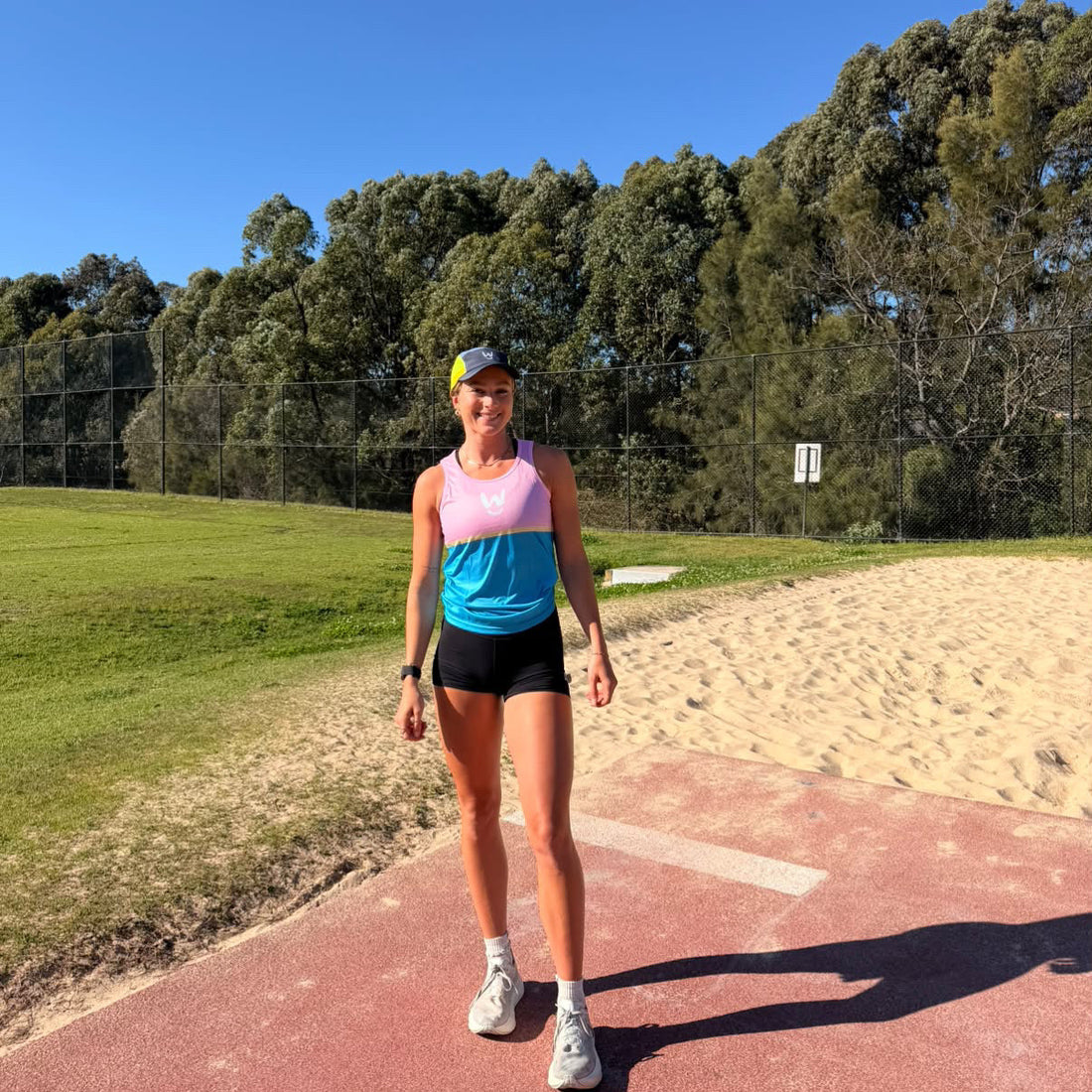 A close-up of the Wippet Performance Running running Singlet that is breathable, sweat wicking, ultra light weight featuring a pink upper section with a white stylized 'W' logo, a thin yellow stripe, and a blue lower section 