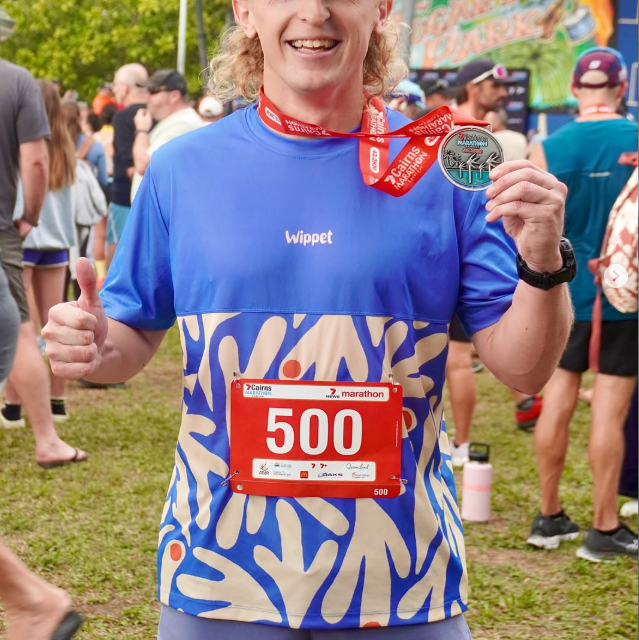 Man wearing Wippet Coconut Cool Everyday Running Shirt with breathable, sweat wicking fabric smiling after race bib and medal, surrounded by people at a running event.